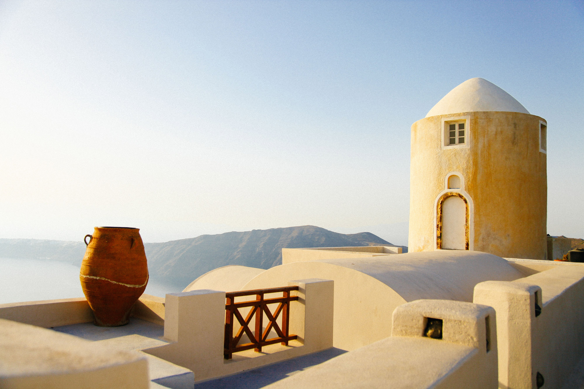 Sunlit whitewashed building with a domed tower and a terracotta urn on a rooftop terrace overlooking a hazy mountainous landscape