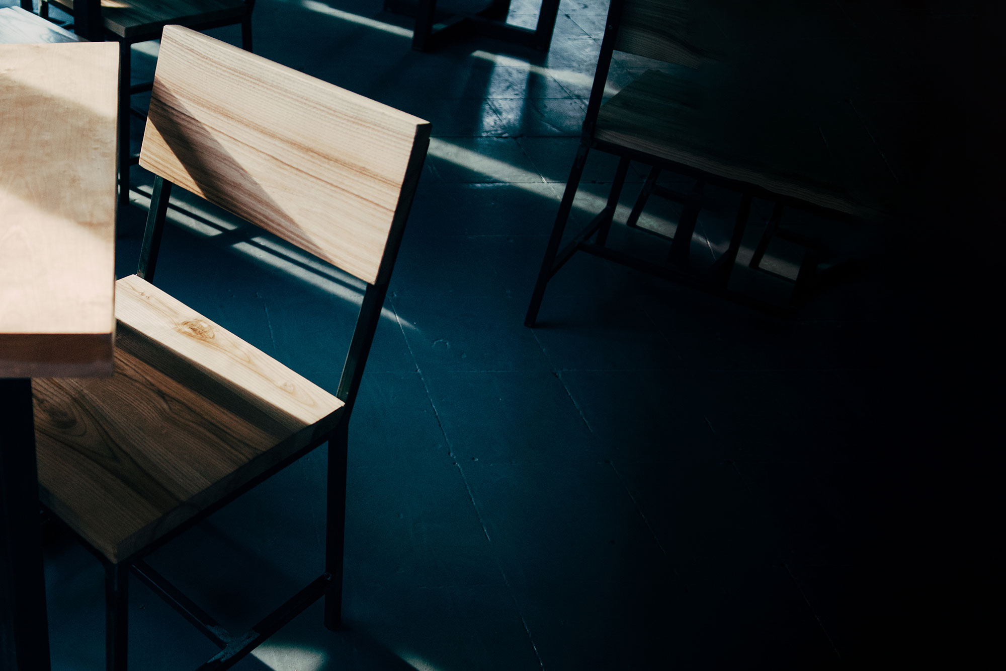Wooden chair and table lit by a diagonal beam of sunlight on a dark teal floor, moody shadowed interior in blue and warm tones
