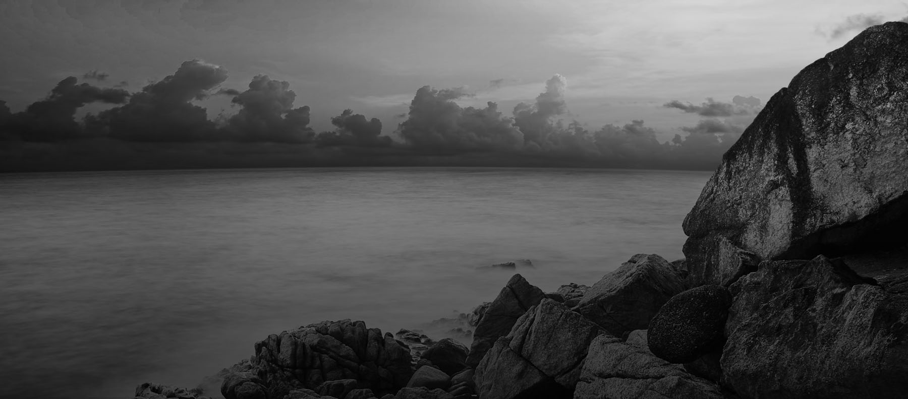 Black and white seascape with large rocks in foreground, calm water, and dramatic cloudy sky at dusk, moody atmosphere